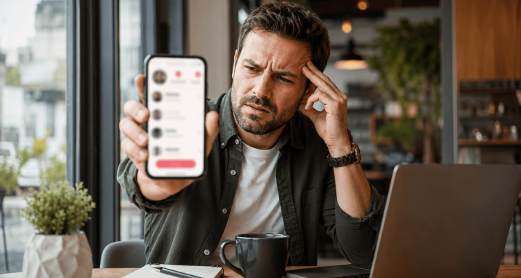 Man in a cafe studies his phone with a puzzled expression, resting his head on one hand while a laptop and coffee sit on the table.