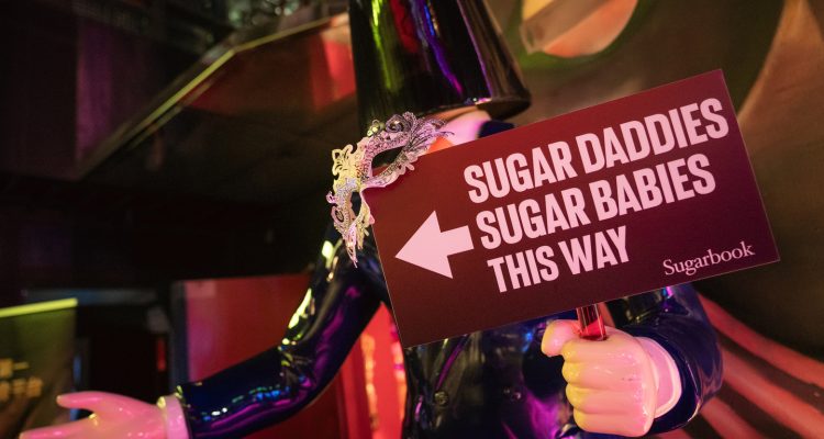 taiwan private party 2026-3 › Sugarbook | The #1 Sugar Daddy & Sugar Baby Dating website to meet Sugar Daddies & Sugar Babies. Build relationships, discuss allowances and get paid instantly. Mannequin in a glossy black outfit holds a maroon sign that says “Sugar Daddies Sugar Babies This Way” with a left-pointing arrow, Sugarbook logo visible.