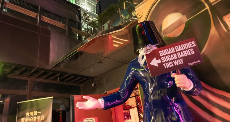 Nighttime city scene with a tall, lit skyscraper in the background and a glossy purple statue in the foreground holding a sign that directs to Sugar Daddies/Sugar Babies.](