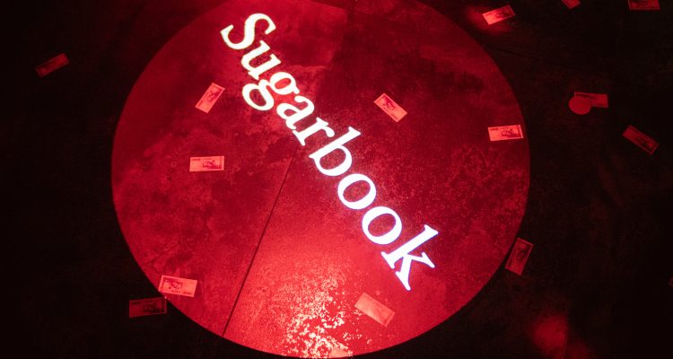taiwan private party 2026-9 › Sugarbook | The #1 Sugar Daddy & Sugar Baby Dating website to meet Sugar Daddies & Sugar Babies. Build relationships, discuss allowances and get paid instantly. Red-lit stage floor with a circular spotlight reading 'Sugarbook' surrounded by scattered bills on the floor.