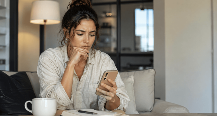 Woman sitting on a sofa looking thoughtfully at her smartphone, with a notebook and pen on the table in front of her.