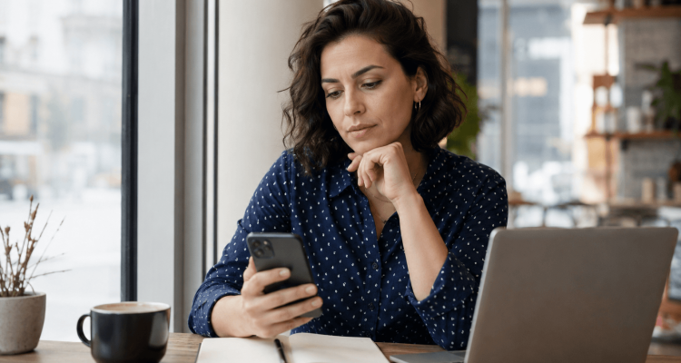 What Should You Check Before Sharing Contact Details › Sugarbook | The #1 Sugar Daddy & Sugar Baby Dating website to meet Sugar Daddies & Sugar Babies. Build relationships, discuss allowances and get paid instantly. Woman in a blue polka-dot shirt sits at a cafe table, looking at her smartphone with a notebook and laptop nearby.