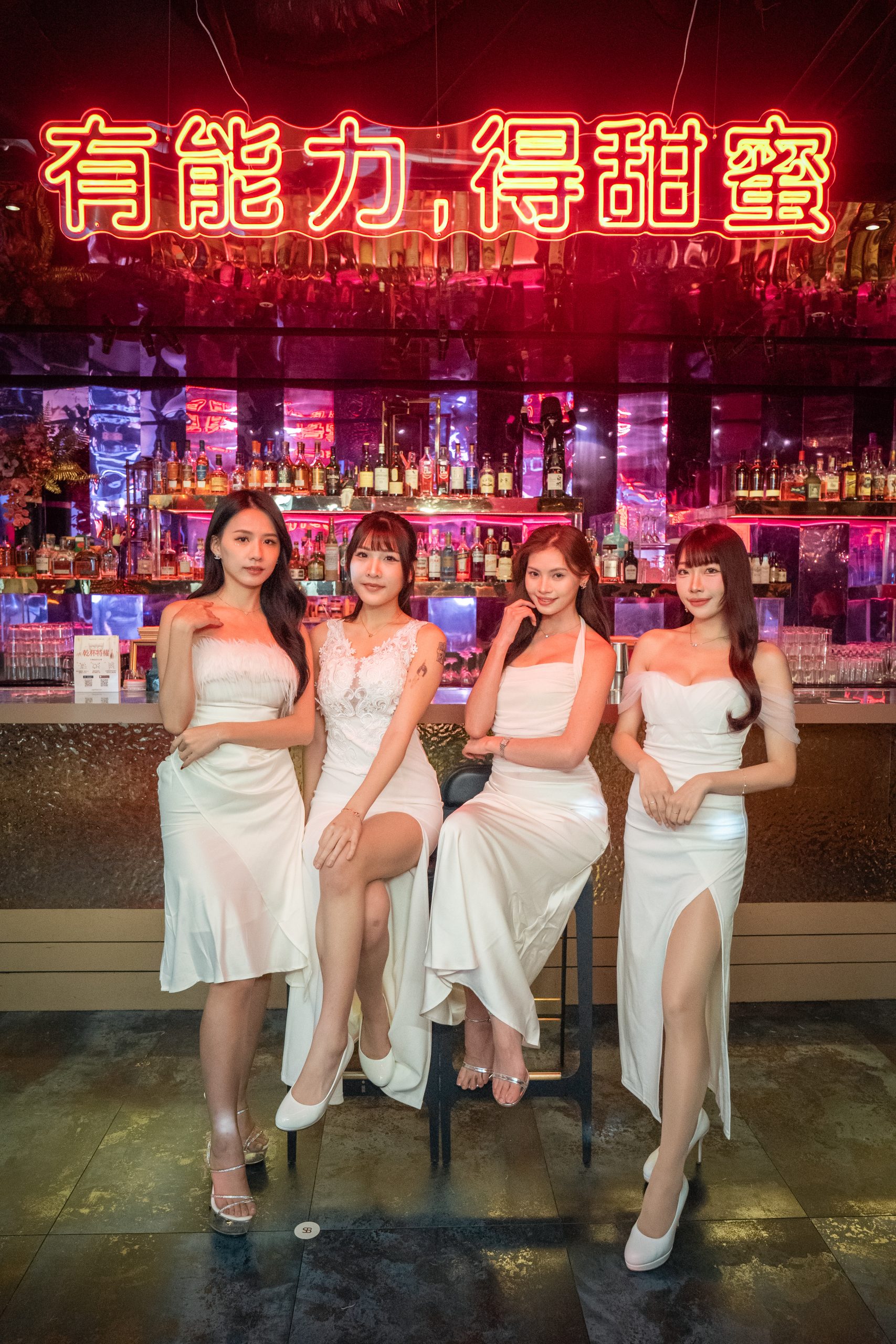 Four women in white dresses pose at a bar with liquor bottles and a red neon sign overhead.