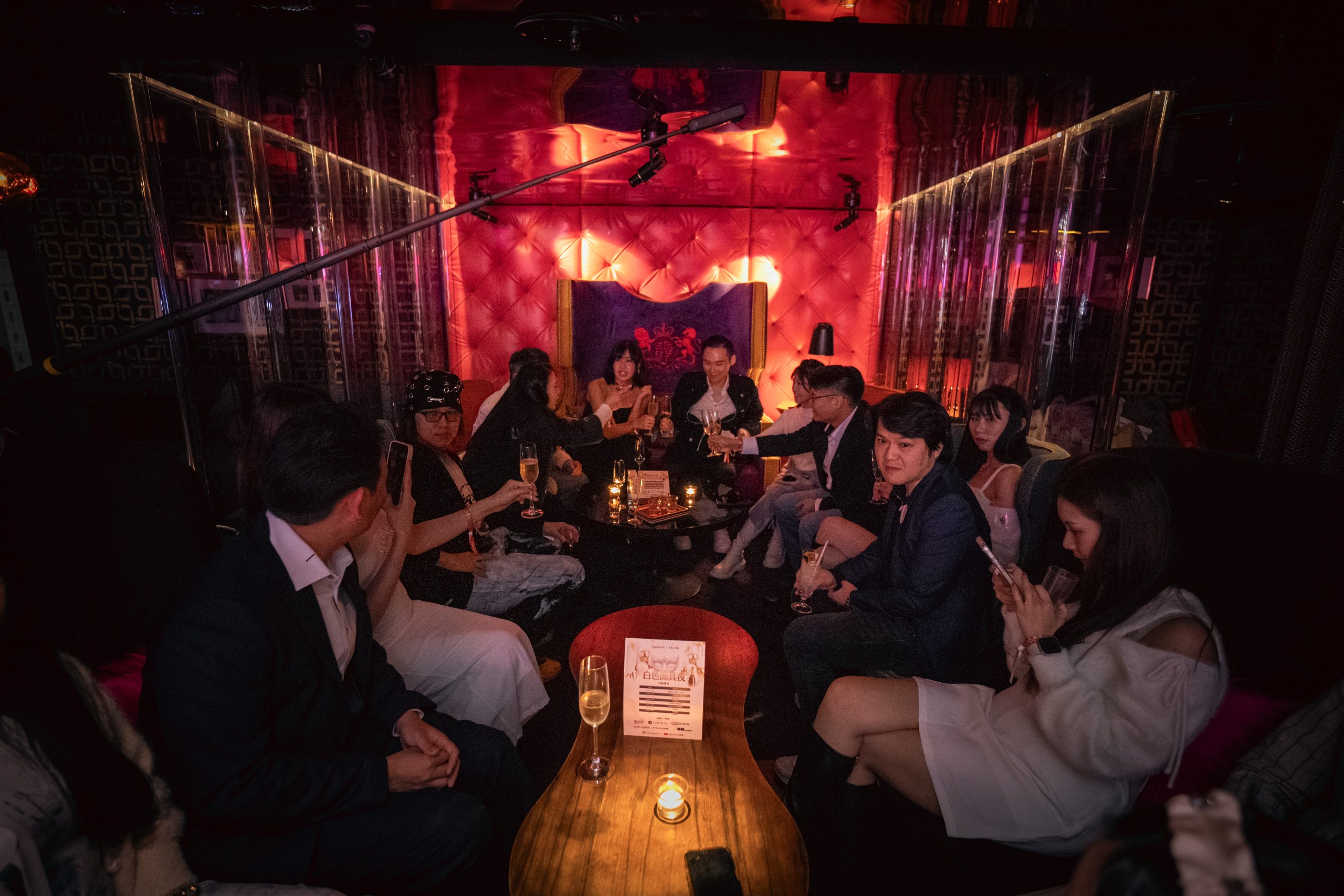 Group of people in a dim club toasting with champagne around a wooden table, red tufted wall behind them and candlelight.",