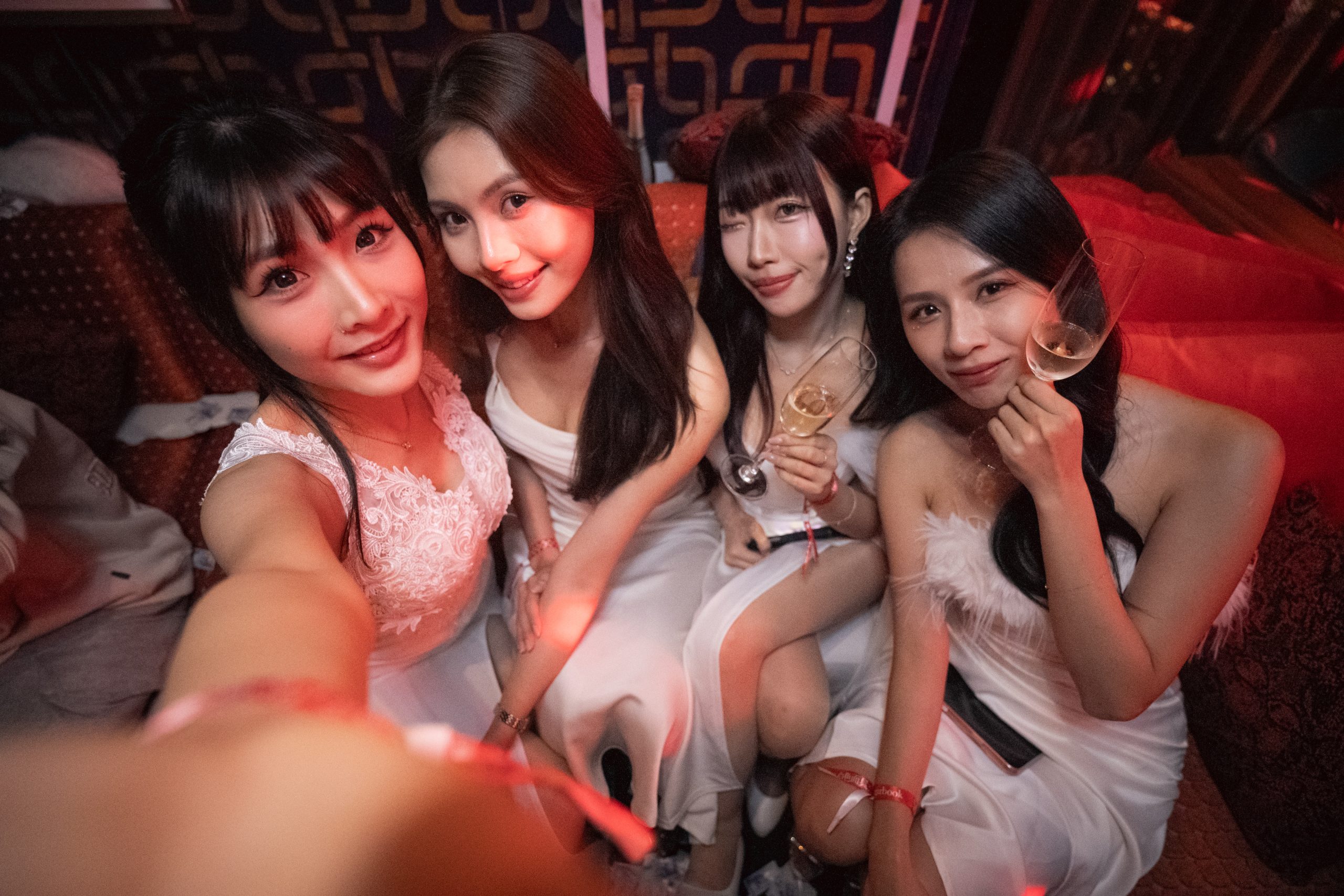 Group of four women in white dresses posing for a selfie at a nightlife venue, holding drinks under red lighting.