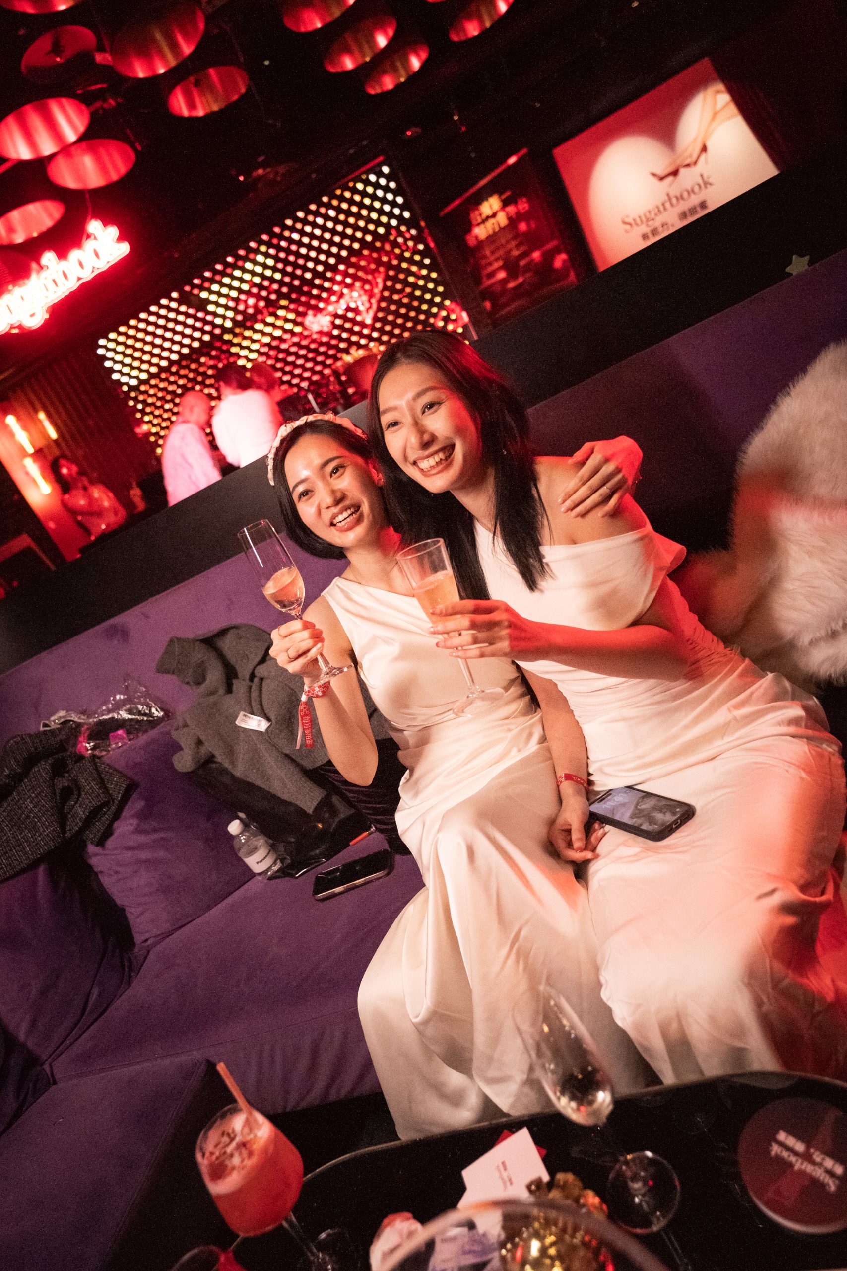 Two women in white dresses posing together, holding champagne glasses in a red-lit club/lounge scene.